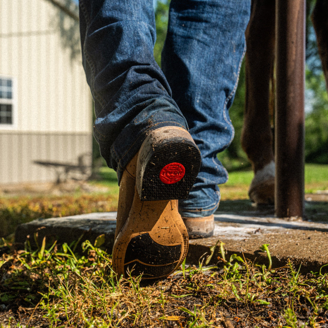 Person wearing blue jeans and brown shoes with a red logo stepping onto grass.