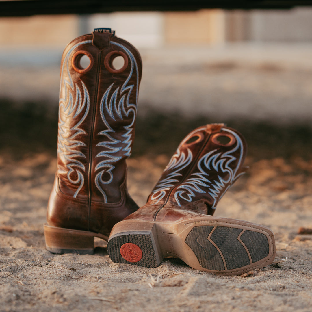 Pair of brown cowboy boots with white embroidery on a dirt ground.