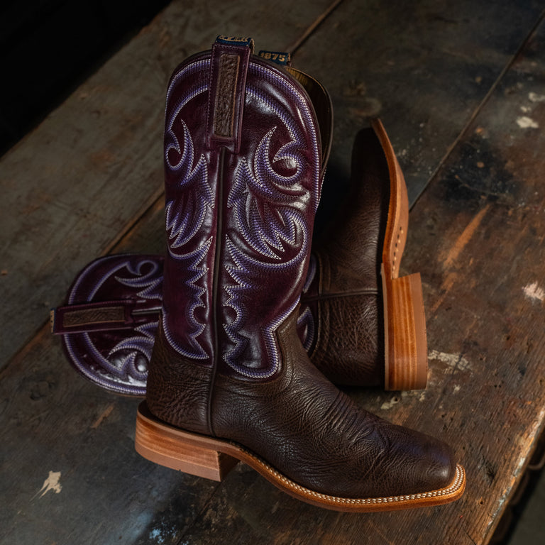 A pair of dark brown leather cowboy boots with purple embroidered shafts, arranged on a rustic wooden surface.