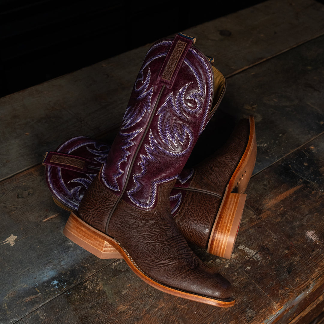 A pair of dark brown leather cowboy boots with purple embroidered shafts, displayed on a rustic dark wooden surface.