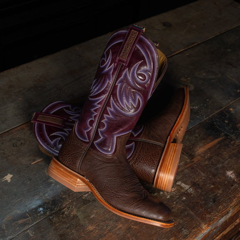 A pair of dark brown leather cowboy boots with purple embroidered shafts, displayed on a rustic dark wooden surface.