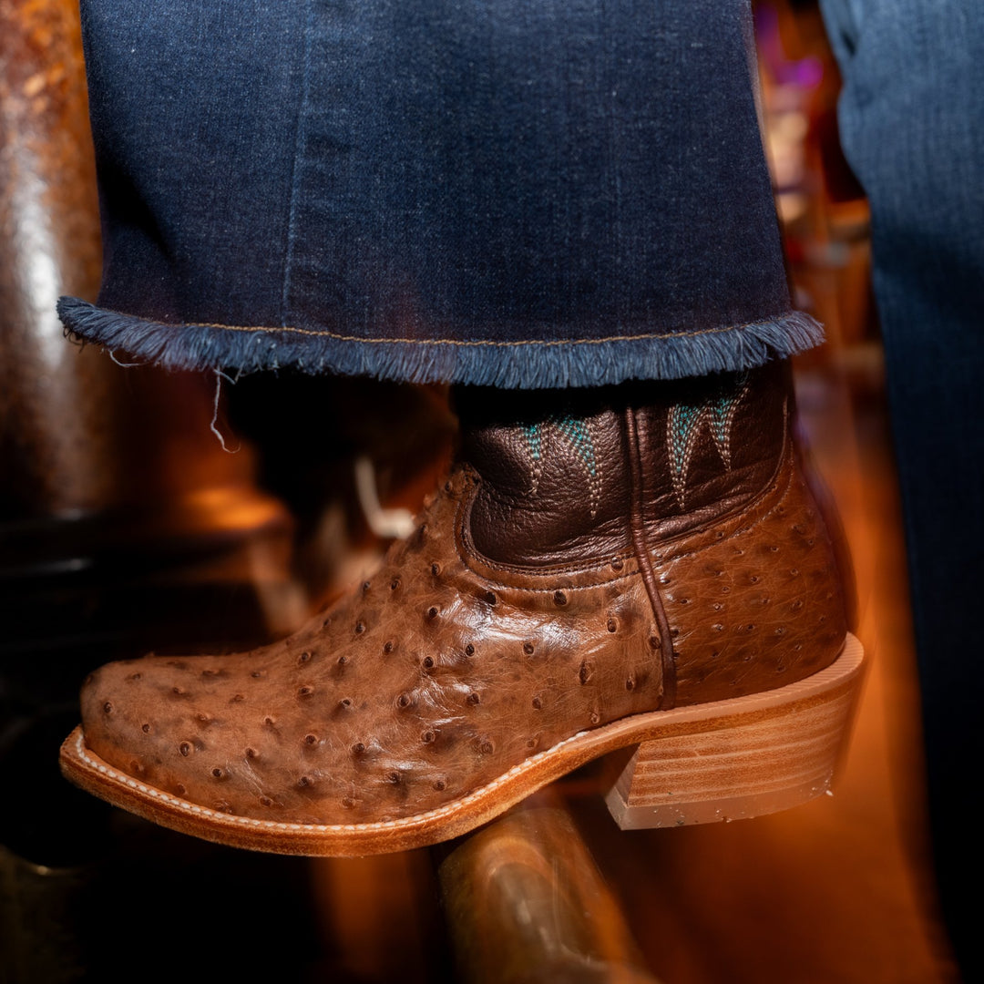 Cowboy boot with brown full quill ostrich vamp and a metallic brown shaft resting on a metal pole under a pair of blue jeans