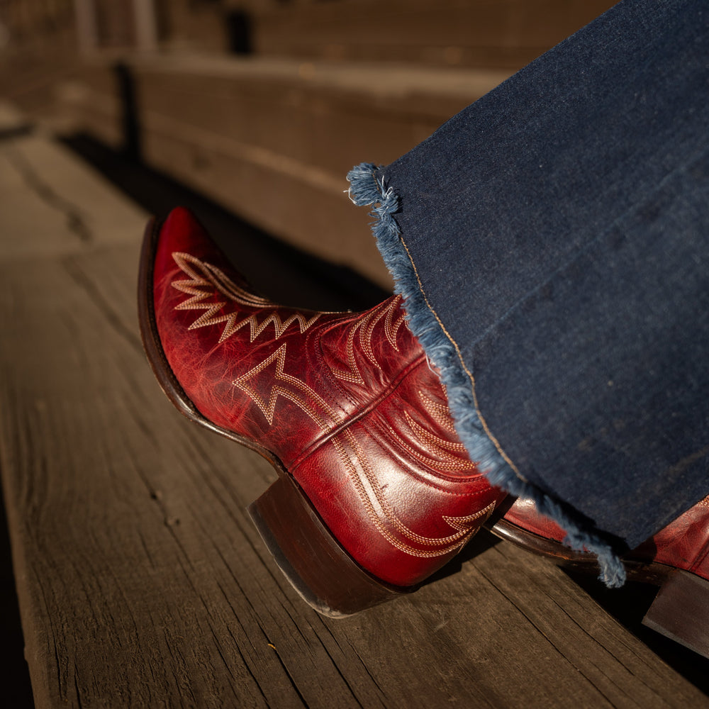 Close up of someone sitting on a set of wooden stairs wearing flared blue jeans and dark red snip toe cowboy boots with contrasting white stitching. 