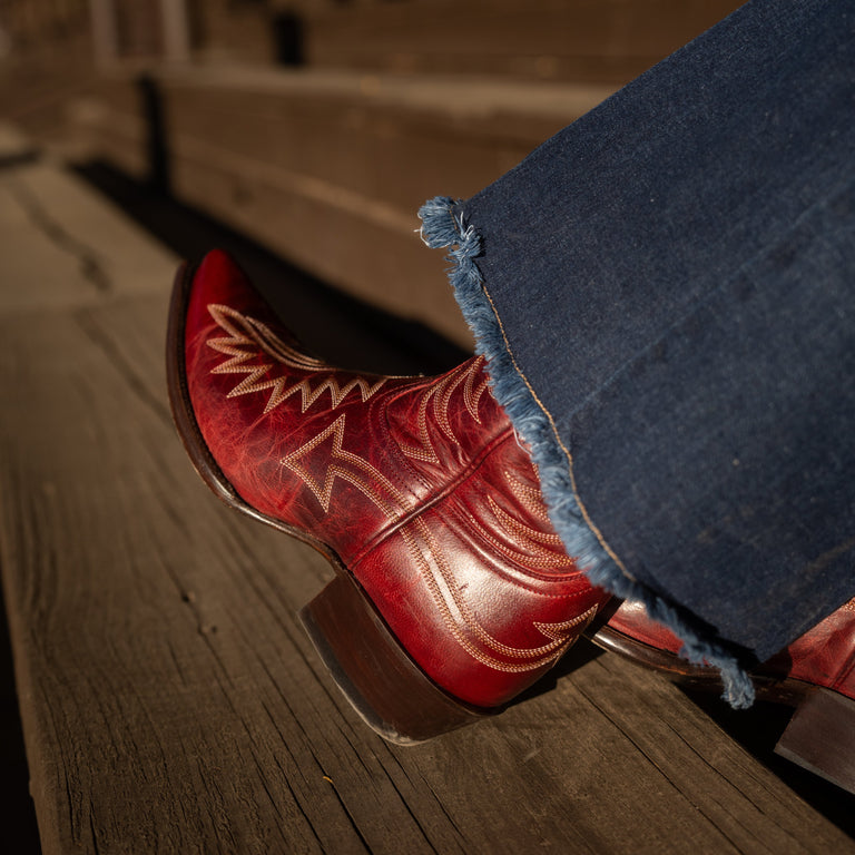 Close up of someone sitting on a set of wooden stairs wearing flared blue jeans and dark red snip toe cowboy boots with contrasting white stitching. 