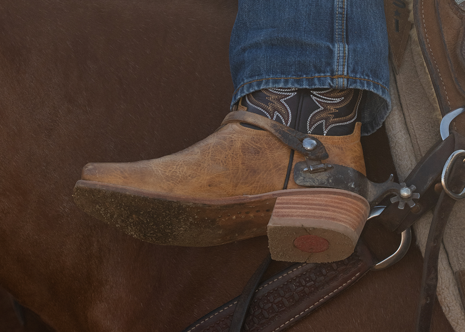 A close-up of a person's foot wearing a brown leather cowboy boot with a stacked leather heel, resting on a horse's footrest with jeans and patterned socks visible.