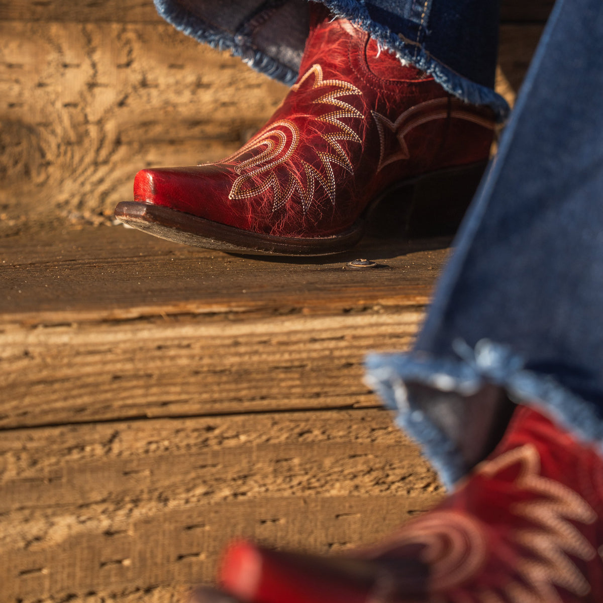 Close up of someone sitting on a set of wooden stairs wearing flared blue jeans and dark red snip toe cowboy boots with contrasting white stitching. 