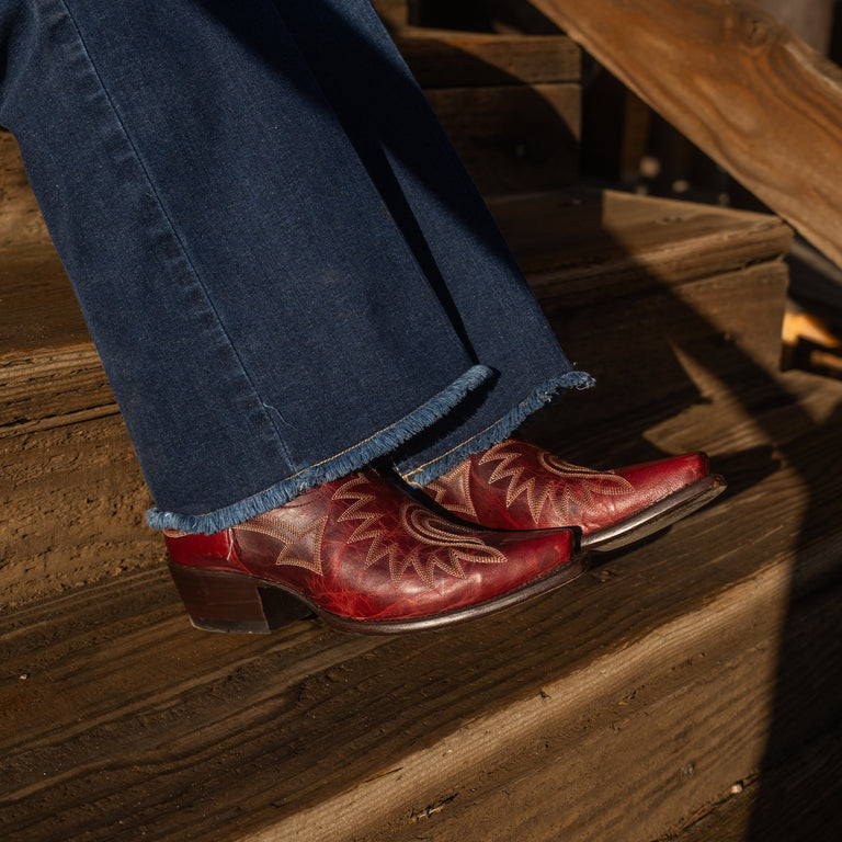 Close up of someone sitting on a set of wooden stairs wearing flared blue jeans and dark red snip toe cowboy boots with contrasting white stitching. 