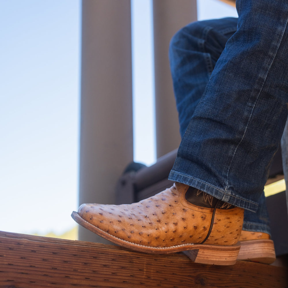 Close up of a person wearing cowboy boots and blue jeans, resting their foot on a wooden surface. The cowboy boots are tan full-quill ostrich.