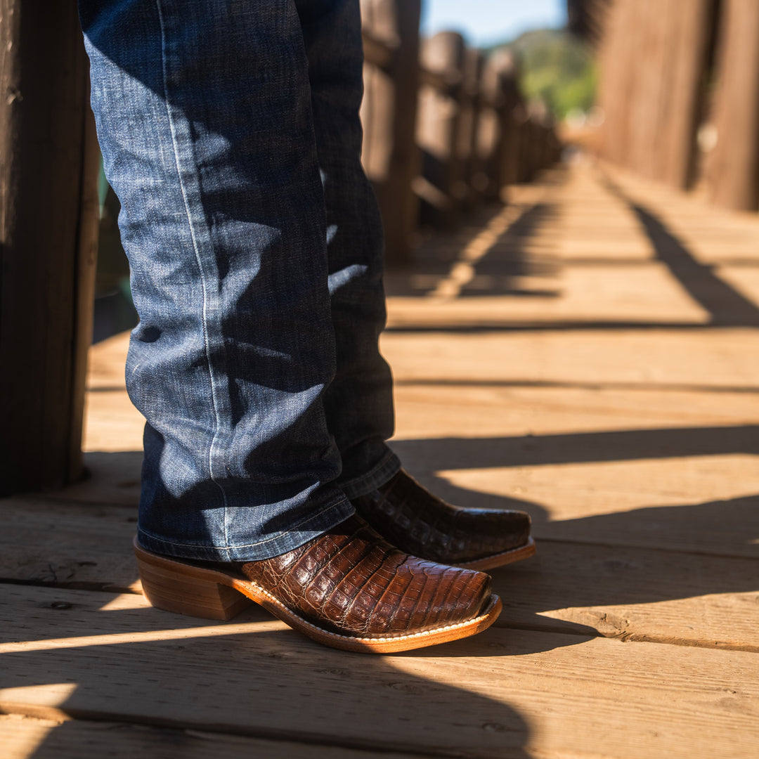 Close up of someone's legs standing on a wooden deck wearing blue jeans and brown caiman cowboy boots.
