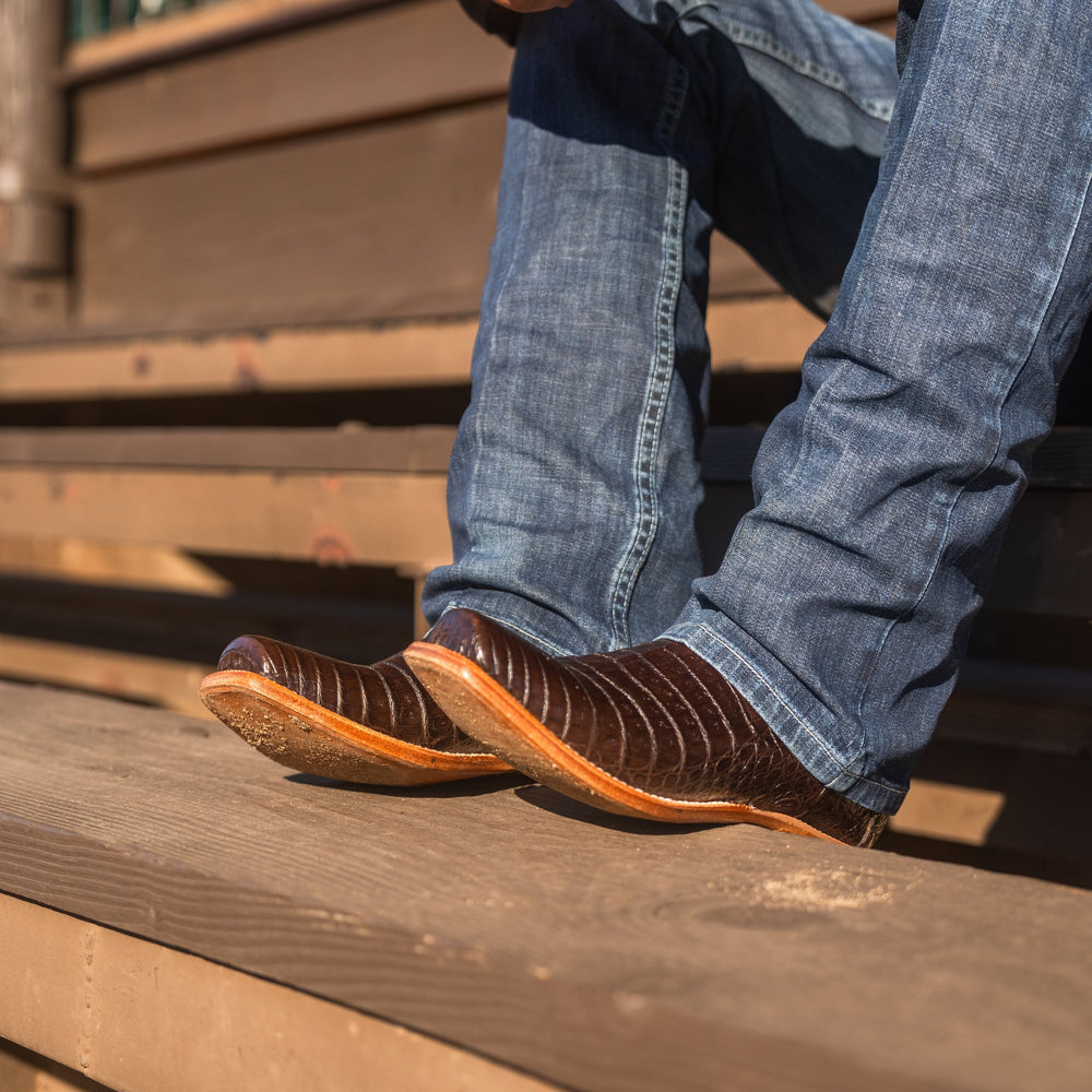 Close up of legs of someone sitting on wooden steps wearing blue jeans and brown caiman cowboy boots