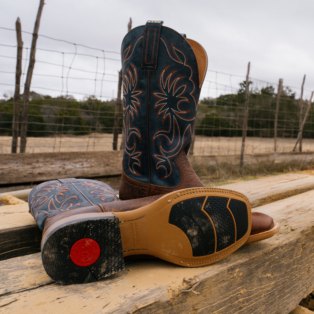 Pair of cowboy boots with intricate designs on a wooden surface, with a fence and trees in the background.