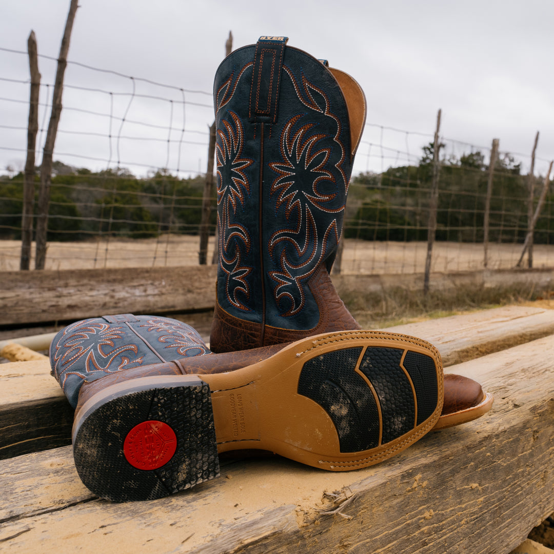 Pair of cowboy boots with intricate designs on a wooden surface, with a fence and trees in the background.