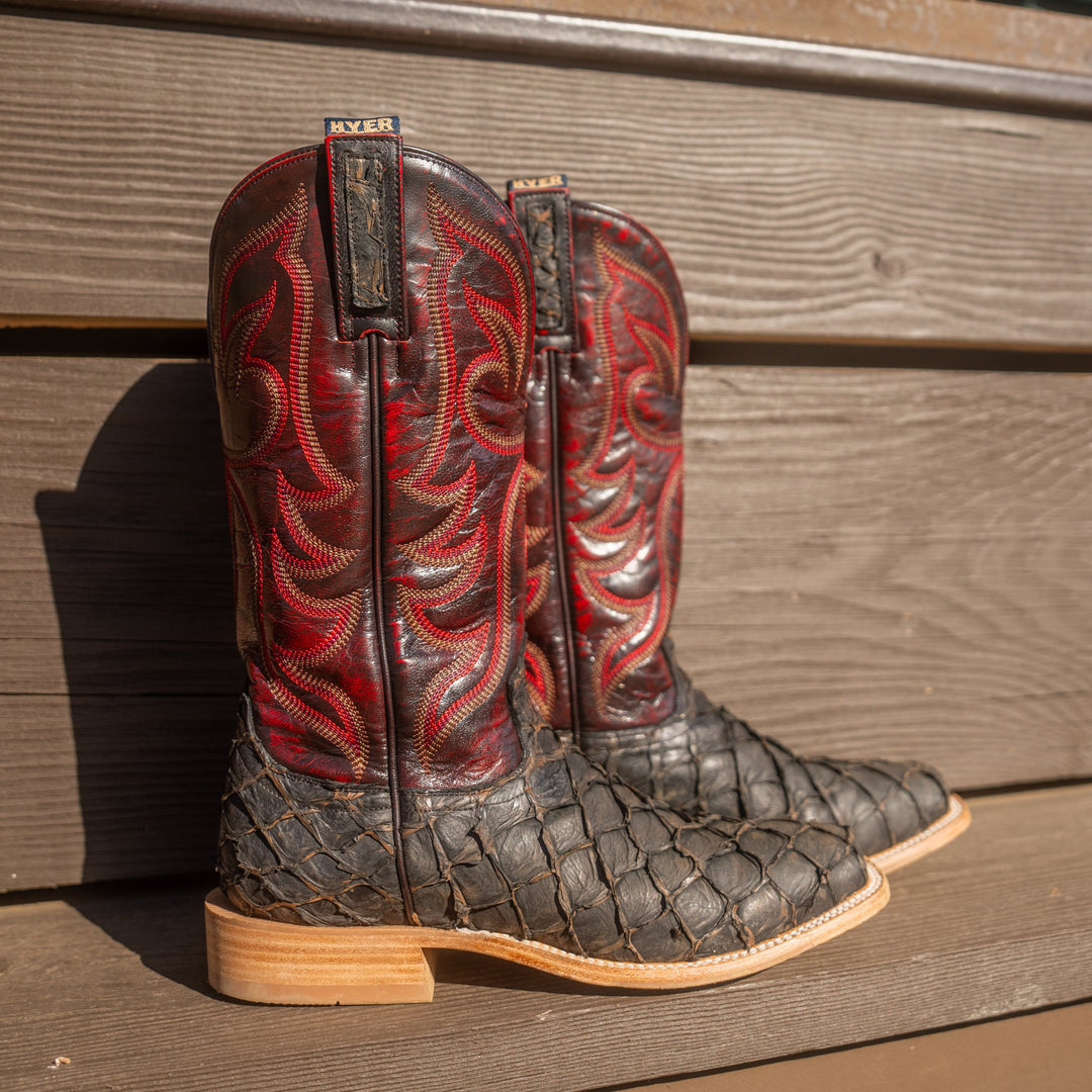 Pair of cowboy boots with dark red shaft with intricate stitching and big bass scaled vamp in front of a wooden slat wall