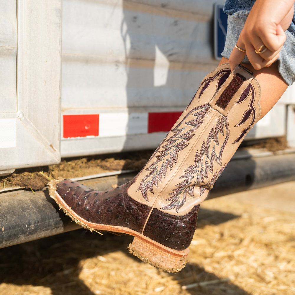 Close-up of a person wearing a purple and beige cowboy boot with intricate designs, standing on a dirt surface.