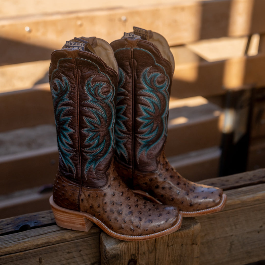 Pair of brown cowboy boots resting on a wooden fence. The cowboy boots have a brown full-quill ostrich vamp, metallic brown shaft and intricate teal stitching.