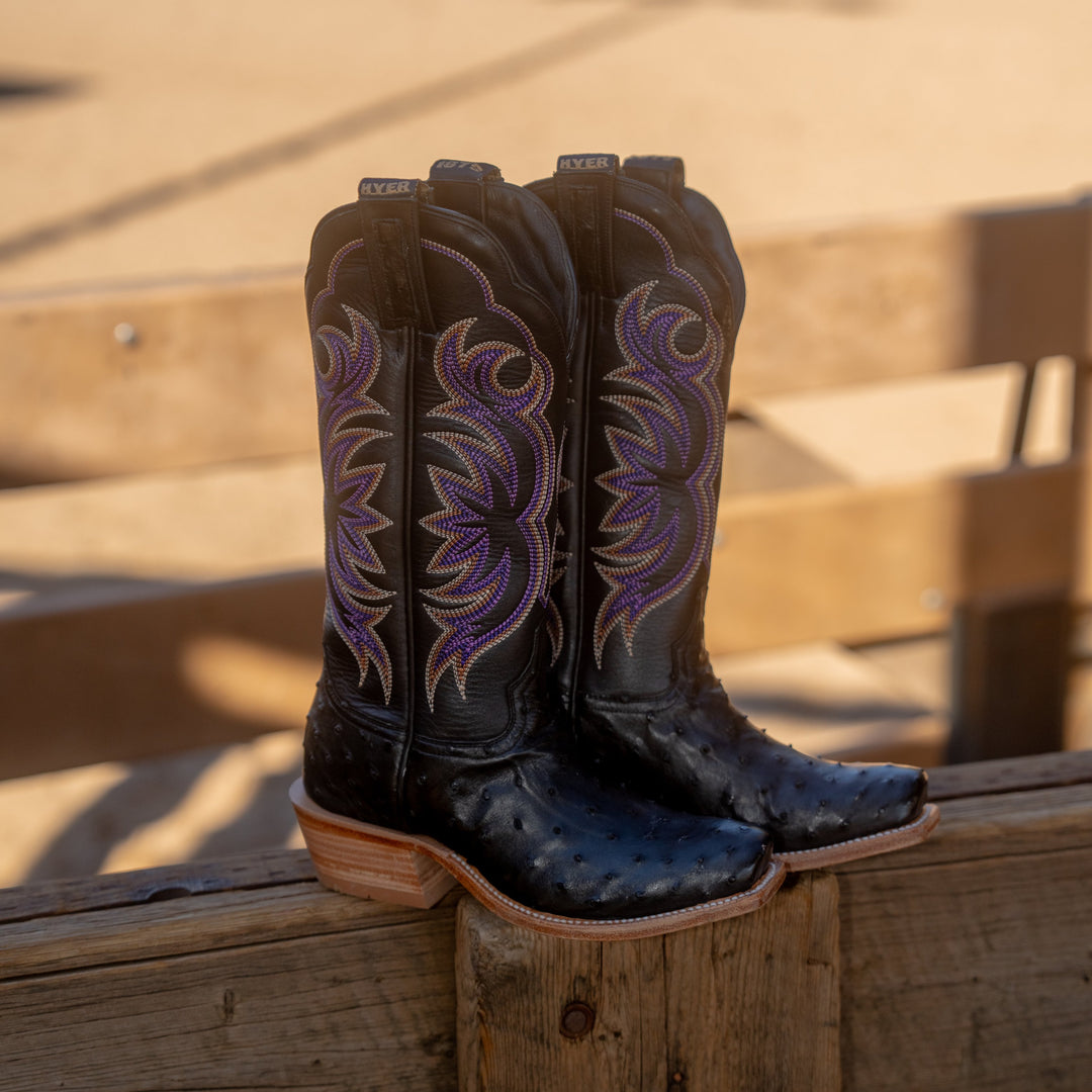 Pair of black cowboy boots with purple stitching and full-quill ostrich vamp sitting  on a wooden slat fence