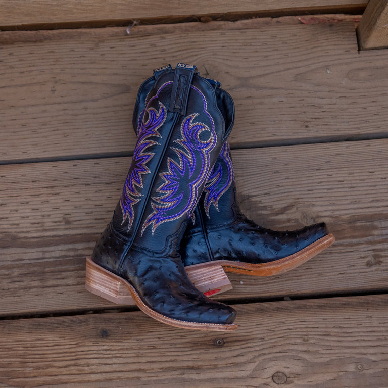 Pair of black cowboy boots with purple stitching and full-quill ostrich vamp laying on a wooden slat floor