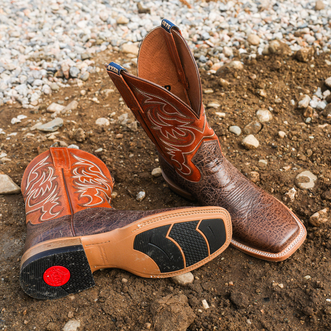 Pair of brown leather cowboy boots with intricate designs on a gravelly ground.