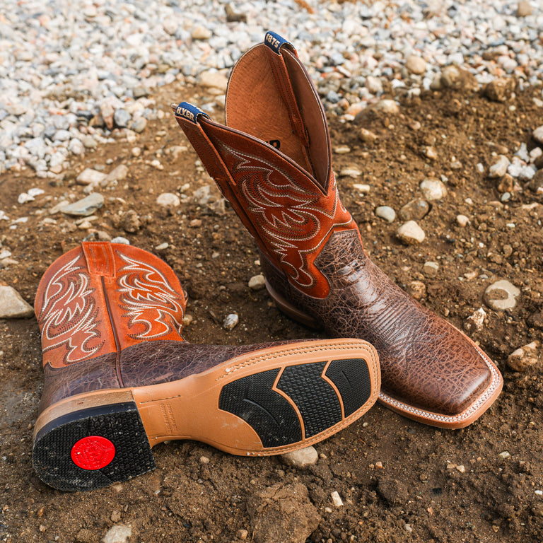 Pair of brown leather cowboy boots with intricate designs on a gravelly ground.