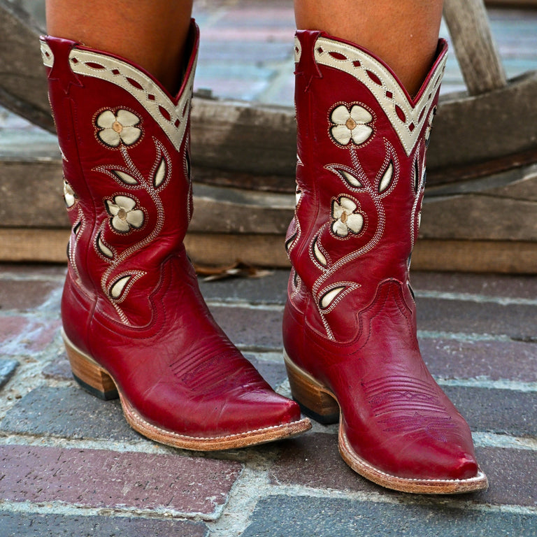 A pair of red leather cowboy boots with cream floral embroidery and western-style stitching, worn on a person's feet on a brick surface with a wooden chair in the background.