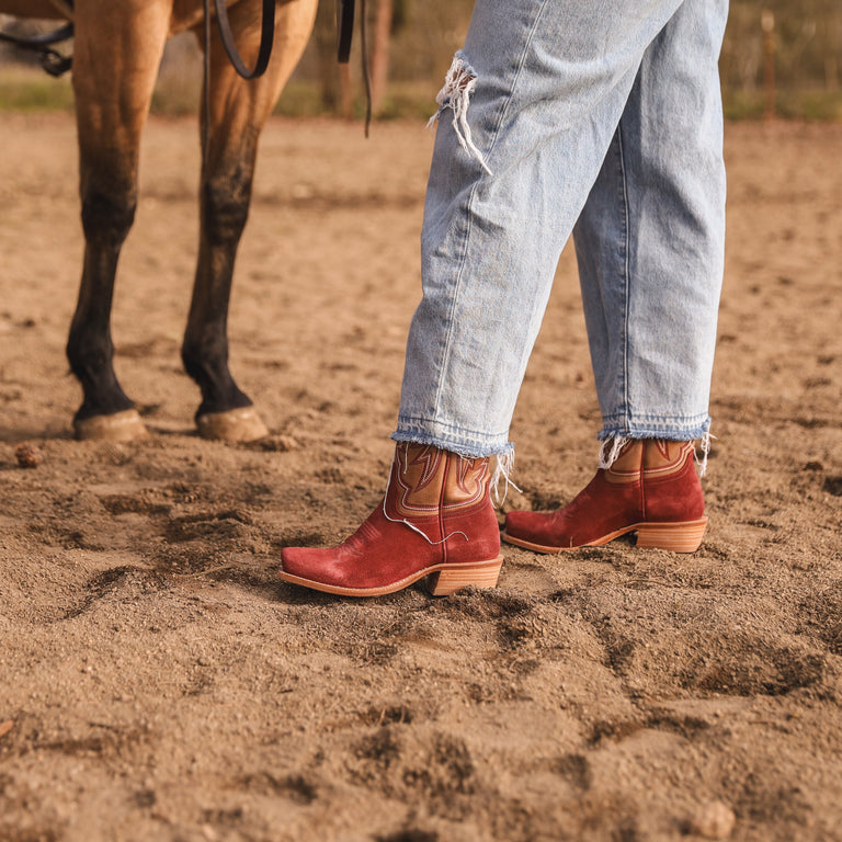 Red cowboy boots and blue jeans on a sandy ground with a horse in the background