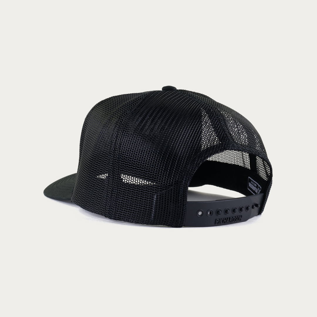 A black trucker-style snapback cap shown from the back, featuring a black mesh back and a solid black flat brim, photographed on a clean off-white background.