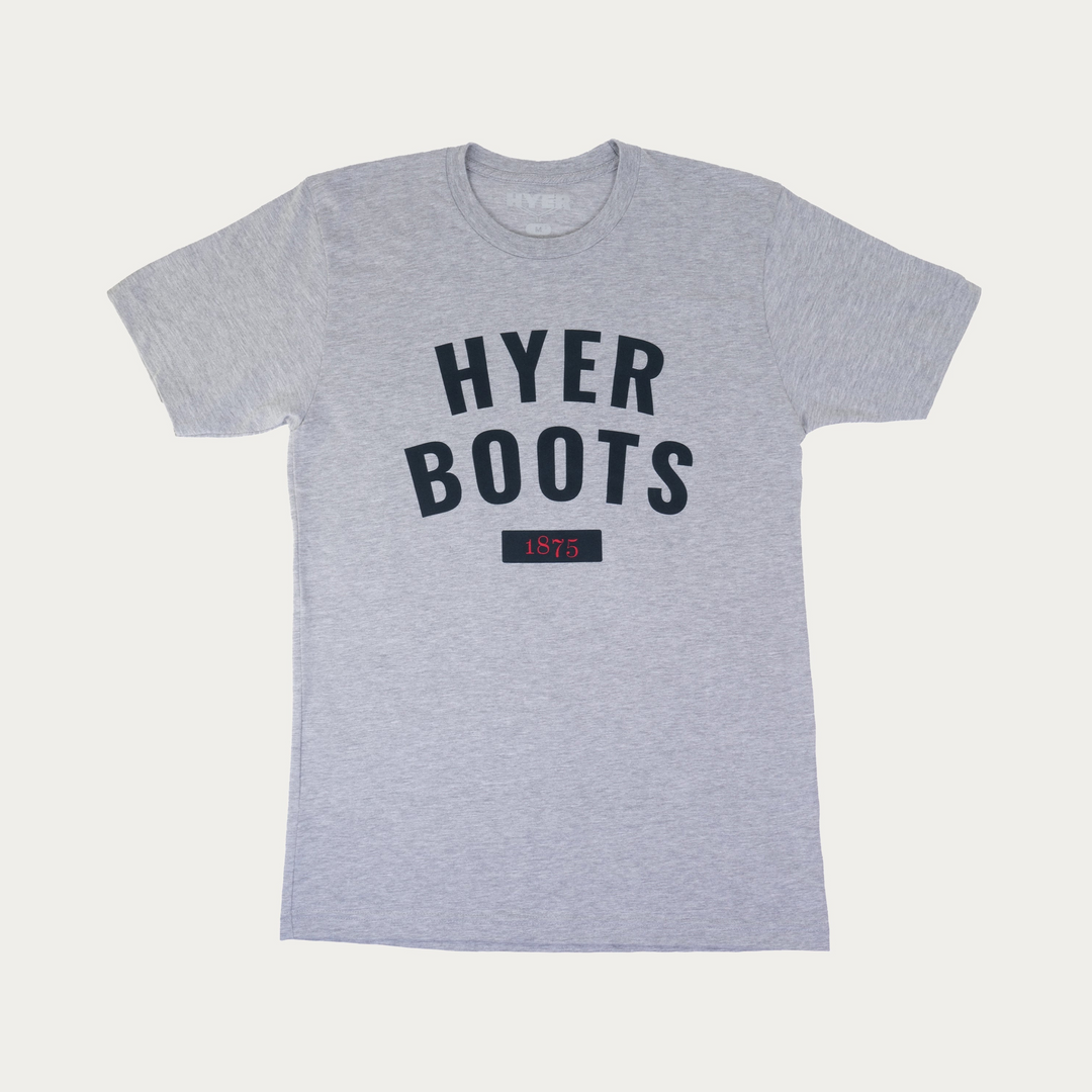 A heather gray crewneck t-shirt featuring the text 'HYER BOOTS' in dark navy blue arched lettering on the chest, with a small dark navy box below containing the year '1875' in red. The shirt is displayed flat against a light off-white background.
