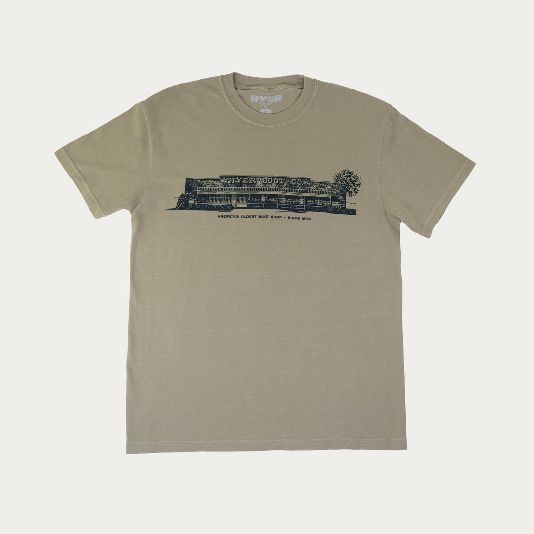 A short-sleeve crewneck t-shirt in a muted khaki-green color, featuring a black graphic print of 'Hyer Boot Co" on the chest, displayed flat against a light off-white background