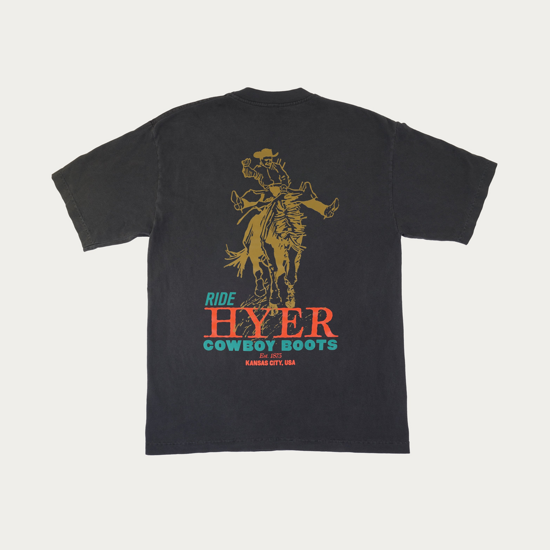 A faded black short-sleeve t-shirt shown from the back, featuring a large graphic print of a cowboy riding a bucking bronco. Below the graphic, the text reads 'RIDE HYER COWBOY BOOTS' in teal and orange-red lettering, with 'Est 1975' and 'KANSAS CITY, USA' printed underneath.
