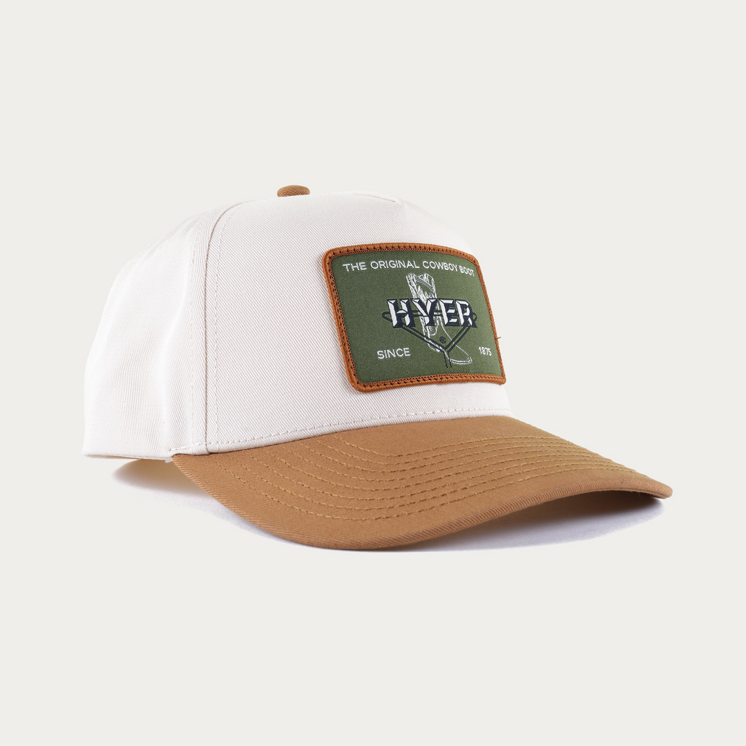 A structured baseball cap with an off-white crown, a tan visor, and a rectangular green patch on the front featuring the brand name 'HYER' and the phrase 'THE ORIGINAL COWBOY BOOT'.