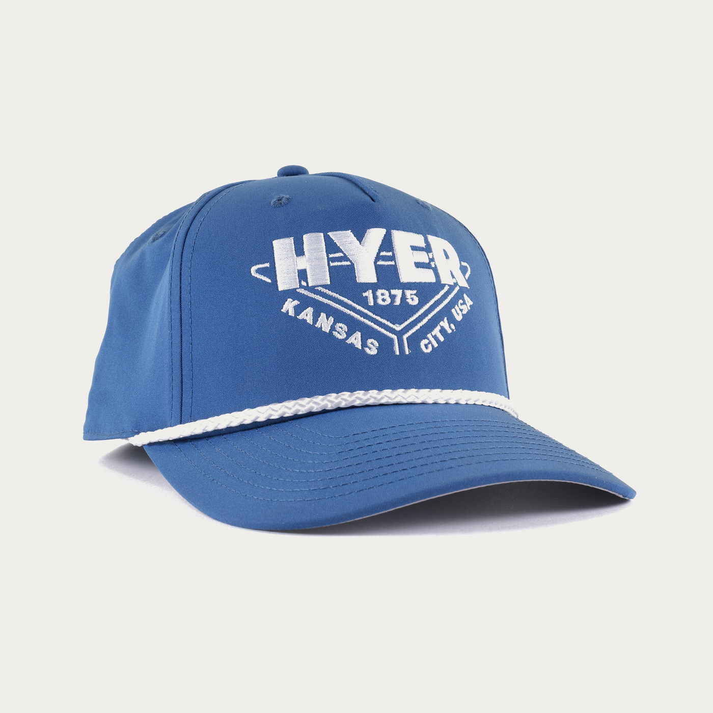 A blue baseball cap with a white rope detail across the brim and a white embroidered graphic on the front reading 'HYER KANSAS CITY, USA' with '1875' in the center, shown on a light gray background.