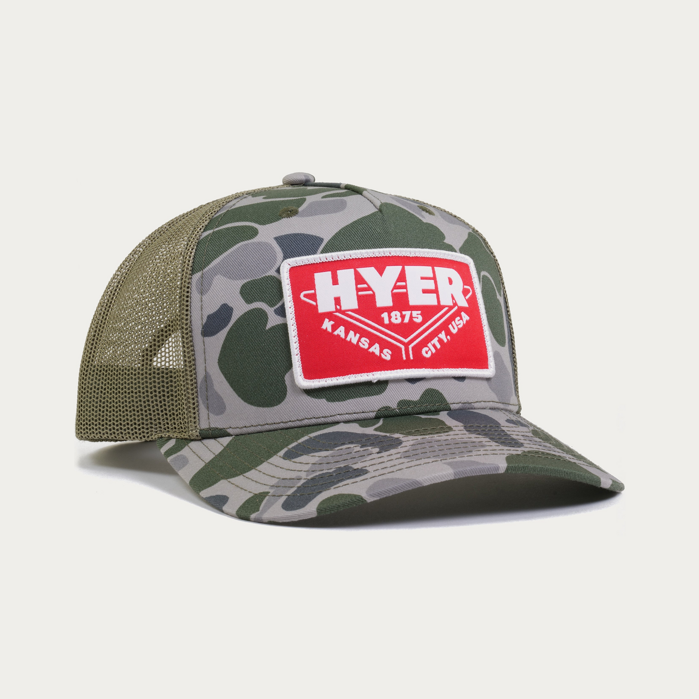 A camouflage trucker hat with green mesh back panels and a red rectangular front patch that reads 'HYER 1875 KANSAS CITY, USA'.