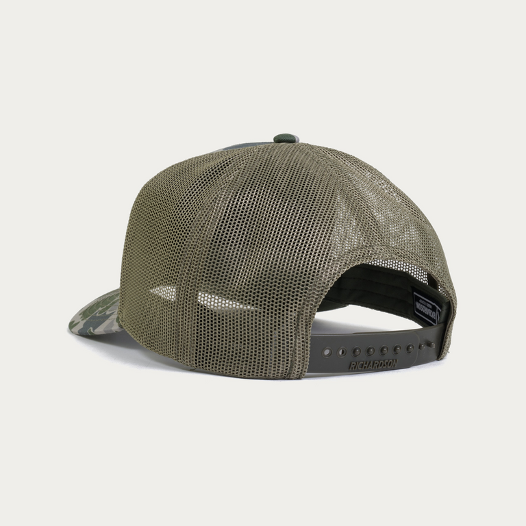 Back view of a trucker-style snapback cap with olive green mesh back panels and a camouflage-patterned visor, on a light gray background.