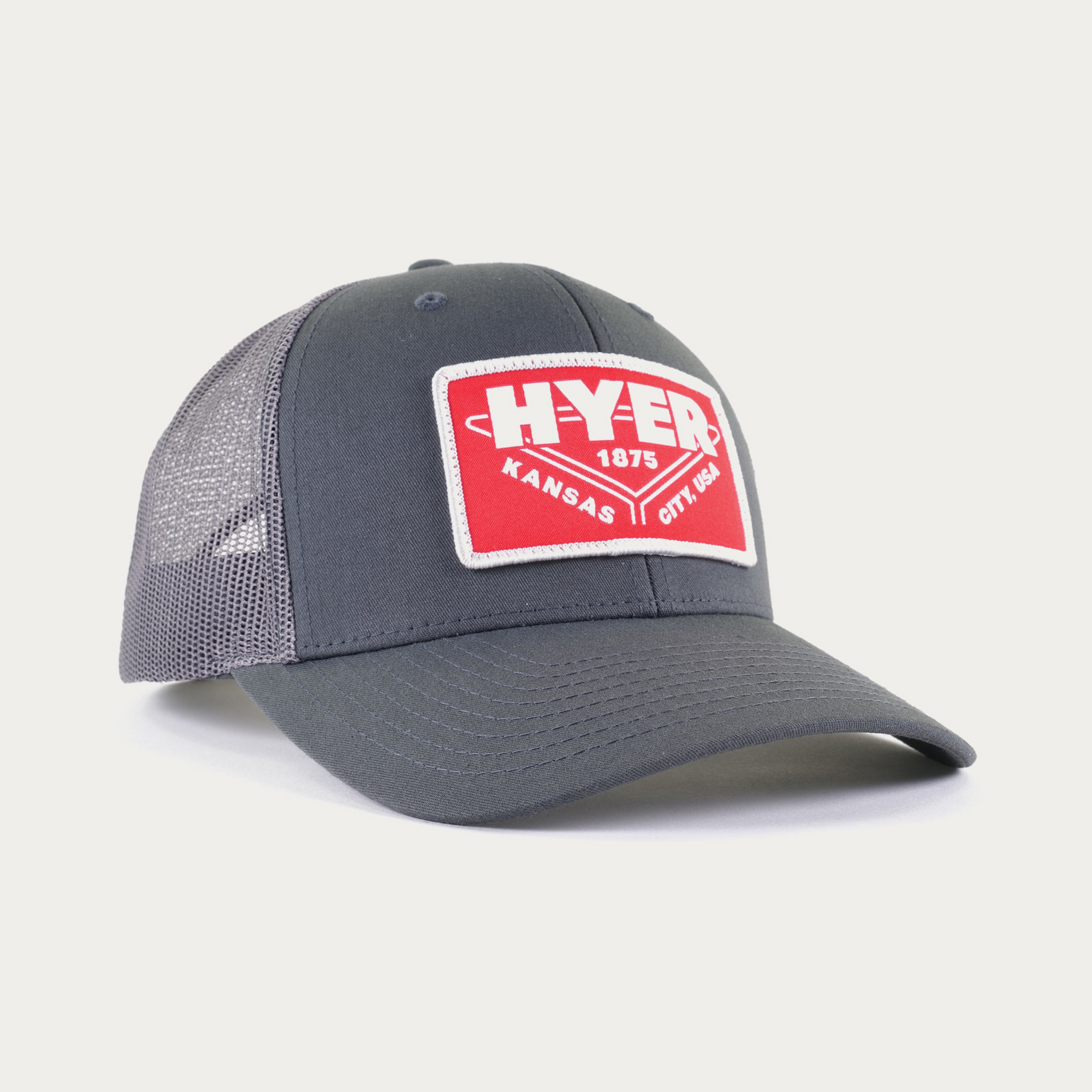 A dark gray trucker hat with a red and white 'HYER KANSAS CITY USA 1875' patch on the front, featuring a gray mesh back, photographed on a light gray background.