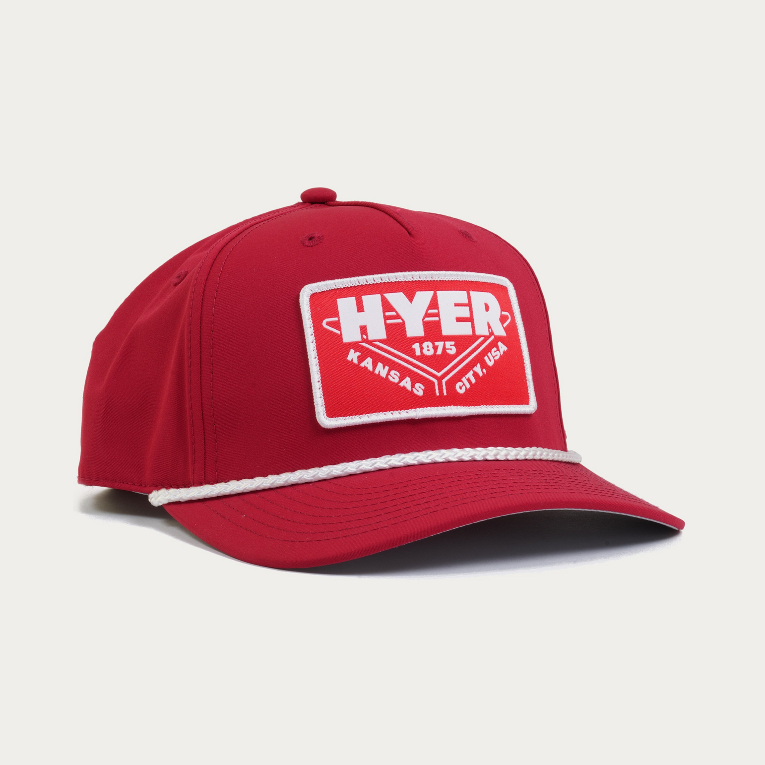 A red baseball cap with a white rope detail along the edge of the brim and a red and white rectangular patch on the front that reads 'HYER KANSAS CITY USA 1875'.