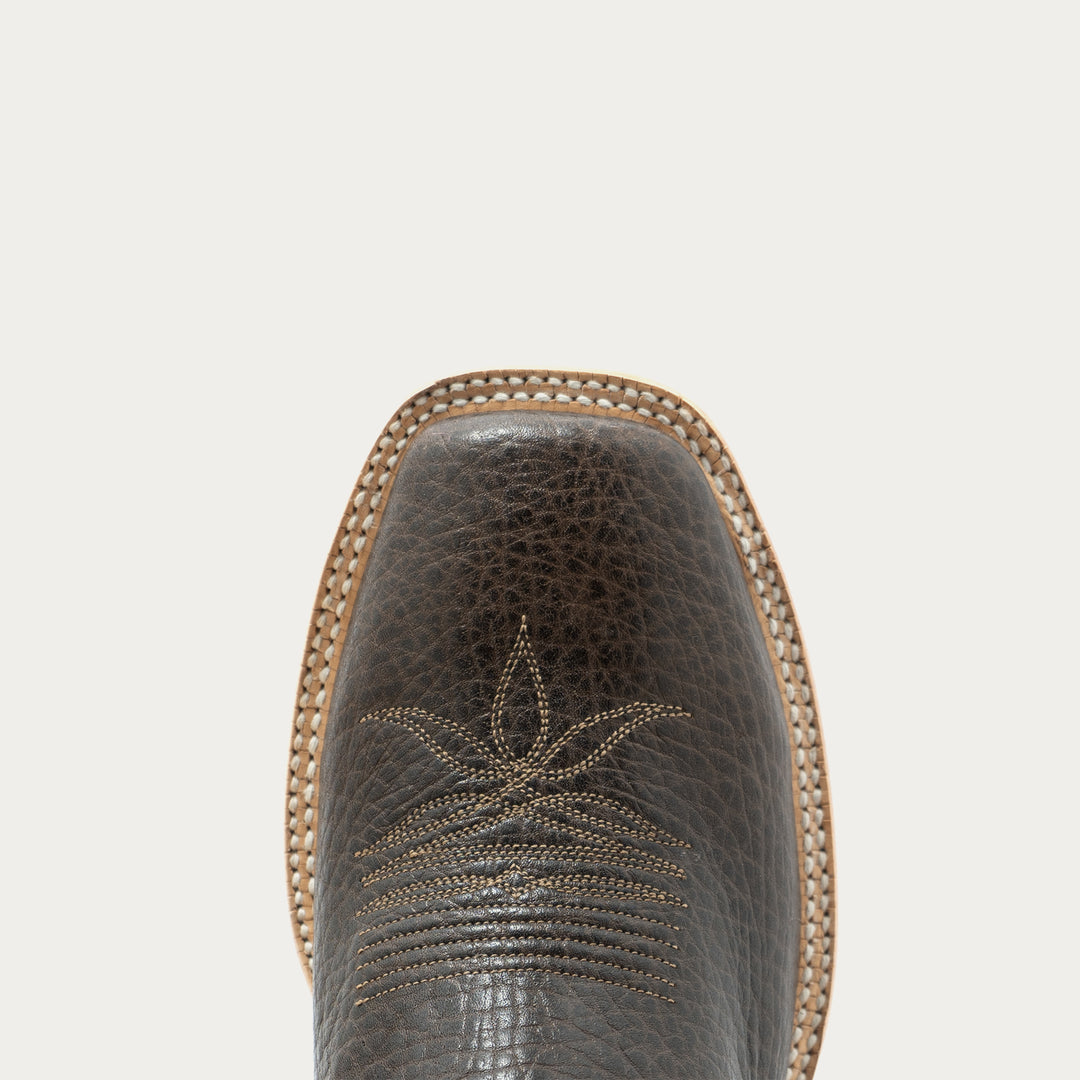 Close-up of the square toe of a dark brown textured leather cowboy boot with decorative stitching, set against a solid off-white background.