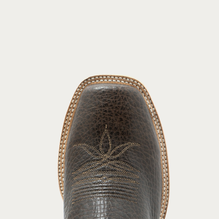 Close-up of the square toe of a dark brown textured leather cowboy boot with decorative stitching, set against a solid off-white background.
