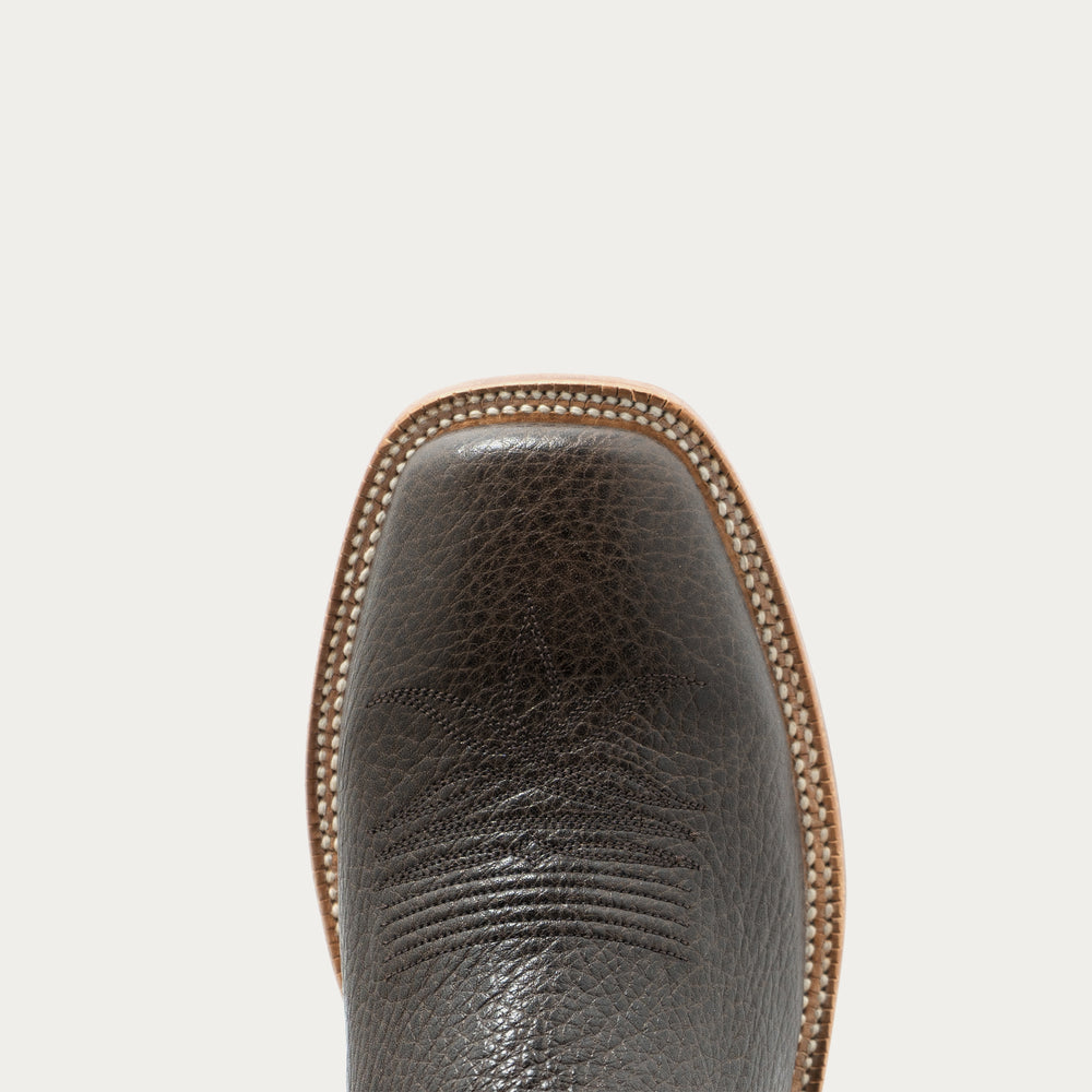 Close-up of the square toe of a dark brown textured leather boot, showing the pebbled leather texture, set against a solid off-white background.