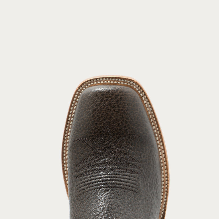 Close-up of the square toe of a dark brown textured leather boot, showing the pebbled leather texture, set against a solid off-white background.