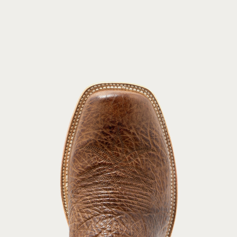 Close-up of the square toe of a brown leather cowboy boot, showing the textured grain and a decorative gold-toned stitched border against a light gray background.
