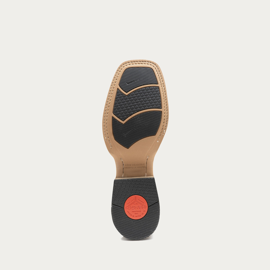 The outsole of a boot is shown from the bottom, featuring a tan rubber footbed, black textured rubber tread, and a small red circular logo on the heel.