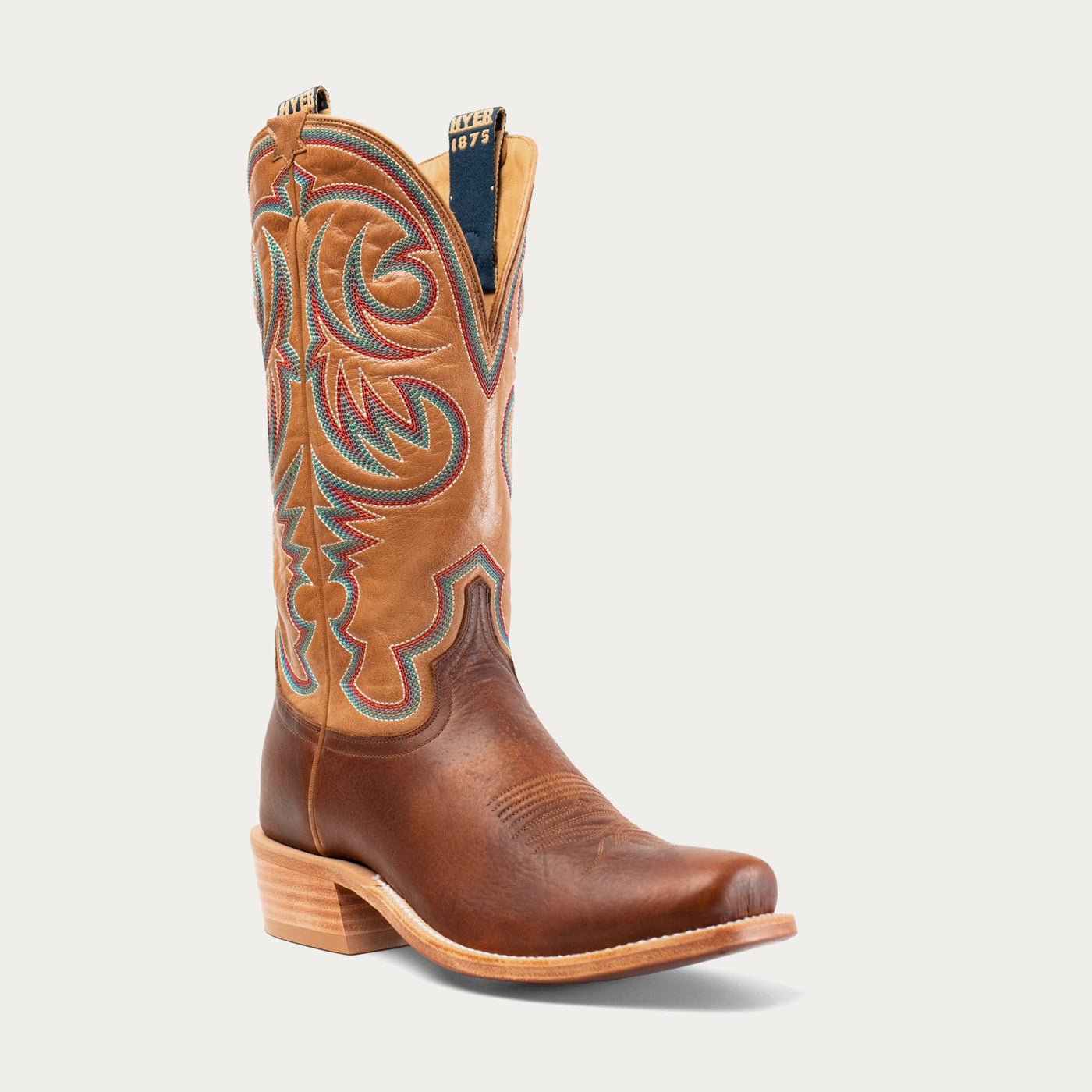 A brown leather cowboy boot with intricate geometric multicolor stitching on the shaft and a wooden heel.