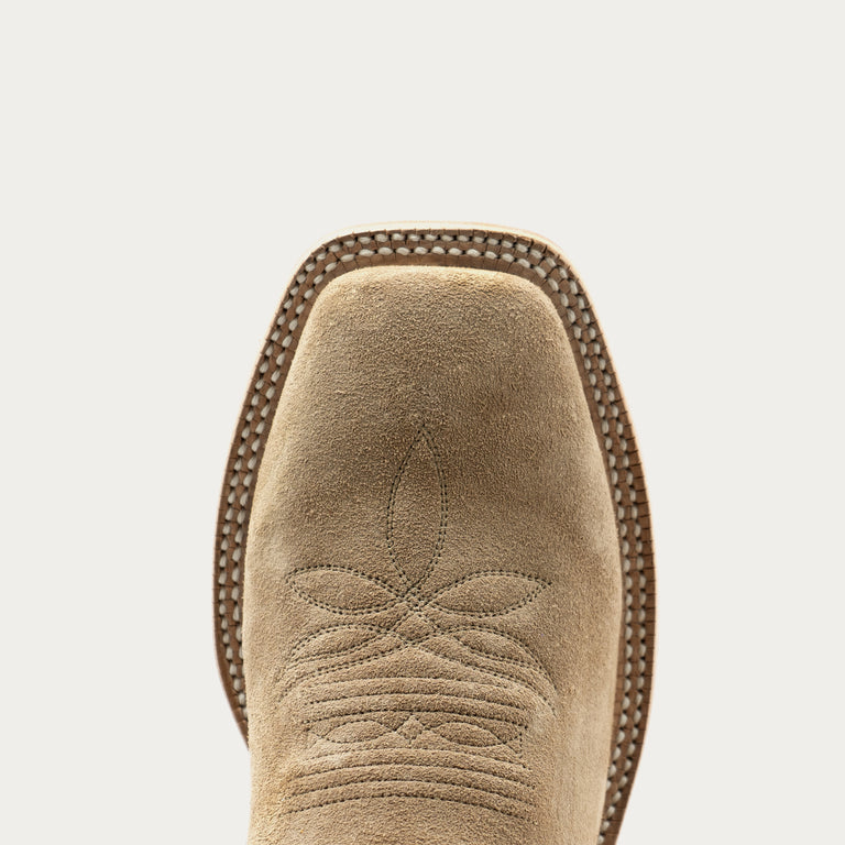 Close-up of the square toe of a light tan suede cowboy boot, showing decorative stitching and a dark brown welted edge against a light gray background.