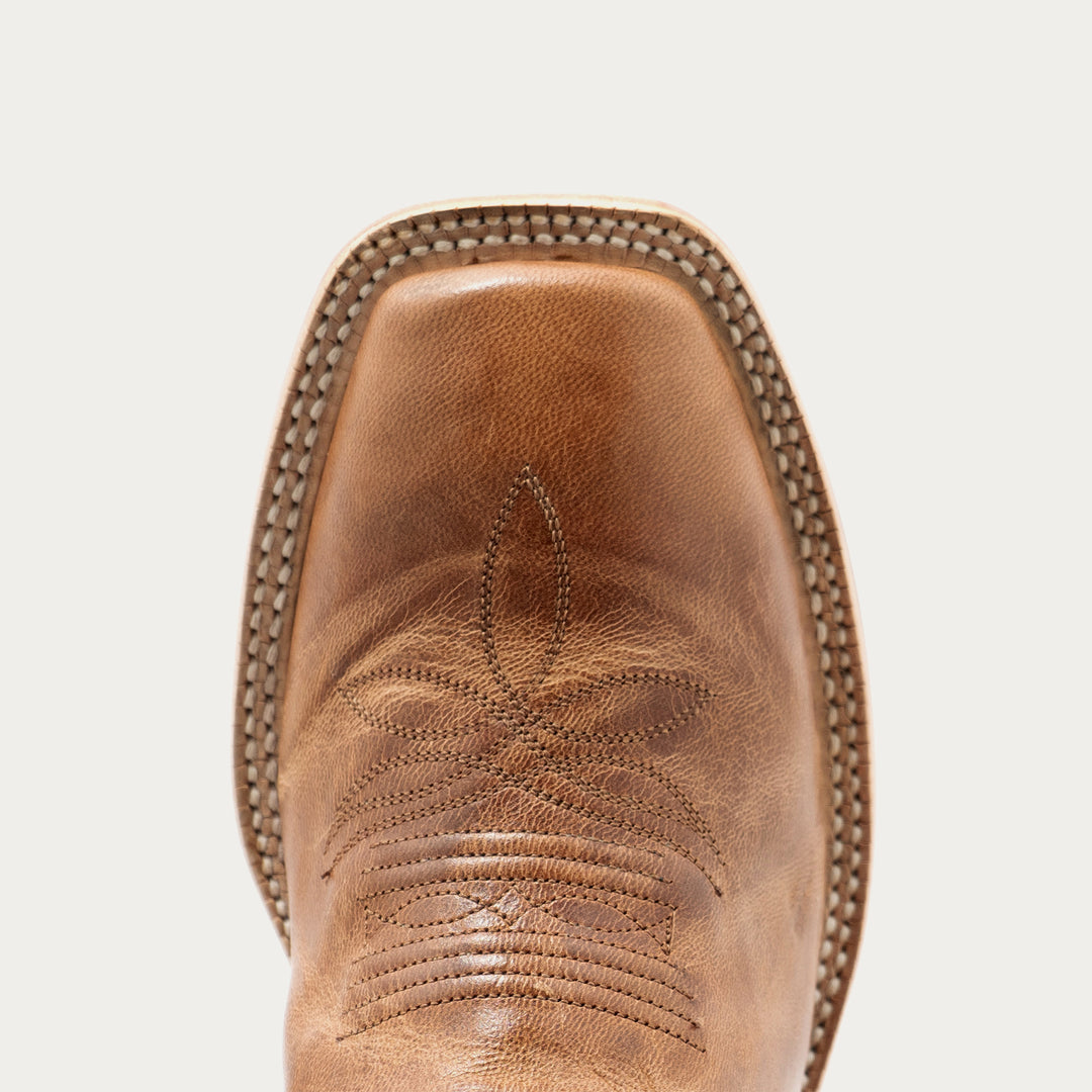 Close-up of the toe of a brown leather cowboy boot with decorative stitching and a pointed square toe, shown against a light gray background.
