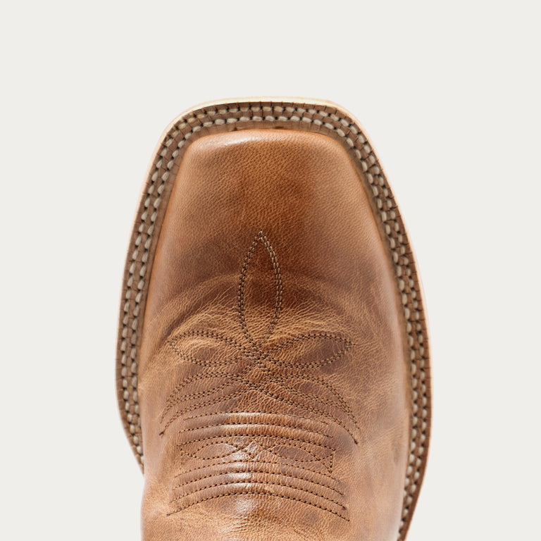 Close-up of the toe of a brown leather cowboy boot with decorative stitching and a pointed square toe, shown against a light gray background.