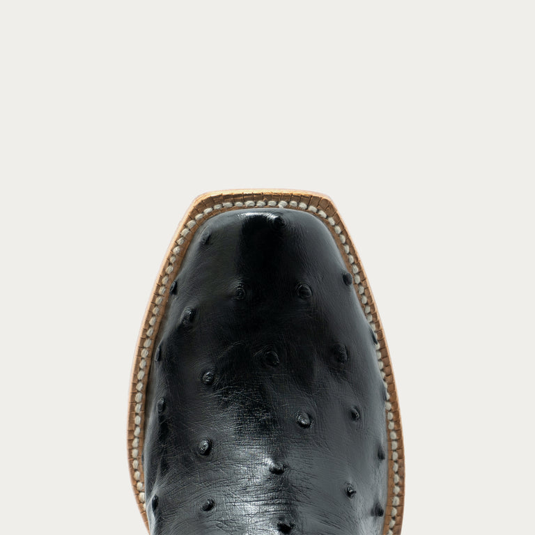 Close-up top view of a black ostrich-textured leather boot with a square toe and decorative tan stitching around the edge, set against a solid off-white background.