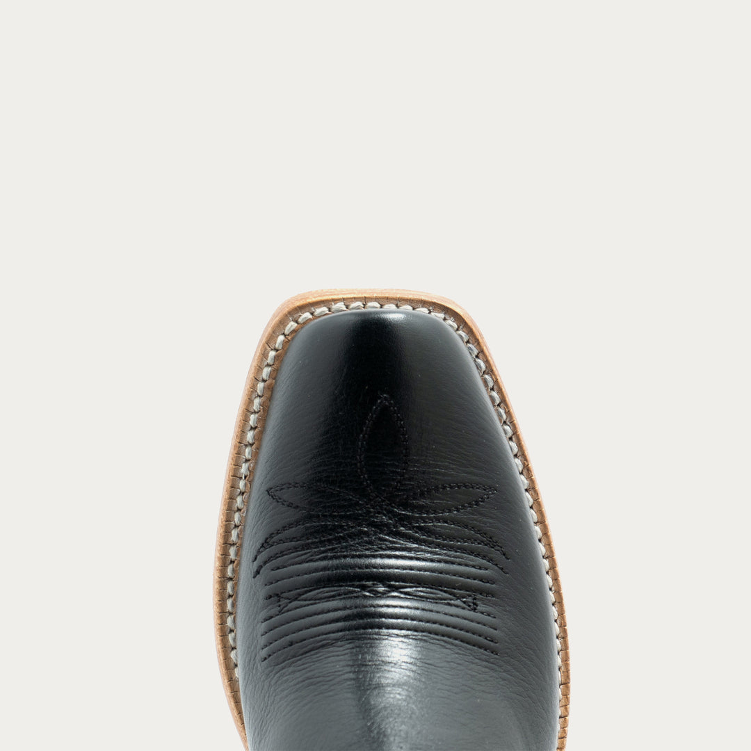 Close-up top view of a black leather cowboy boot toe with decorative stitching, centered on a plain light gray background.