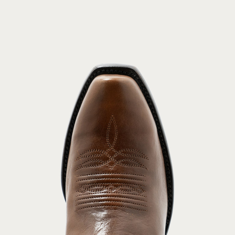 Close up of tan rounded point cowboy boot with tonal stitching on a gray background