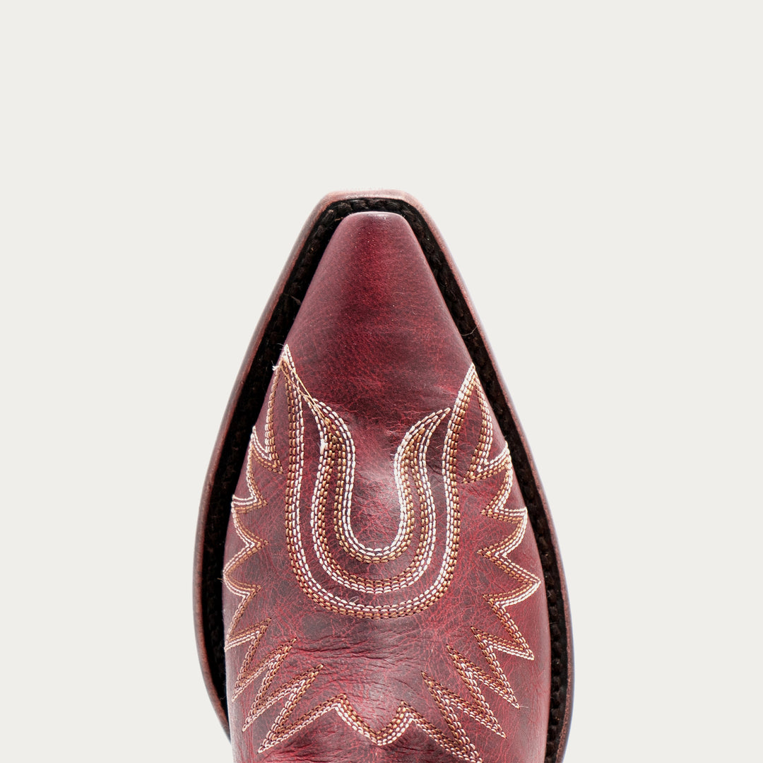 Close-up of a pointed western boot toe in dark red leather with decorative light tan stitching, shown against a light gray background.
