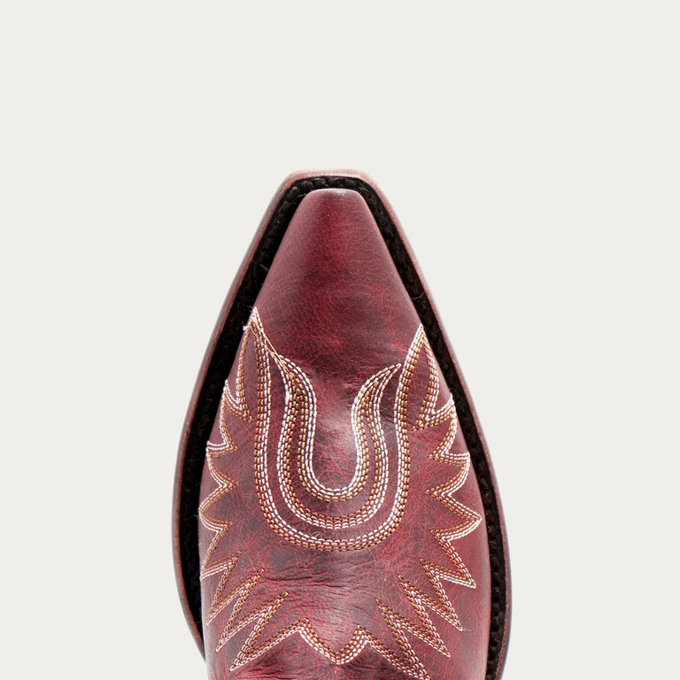 Close-up of a pointed western boot toe in dark red leather with decorative light tan stitching, shown against a light gray background.
